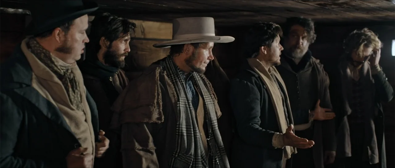 Out of Liberty (2019) movie still: medium — A dark wooden room constructed of logs features a group of men standing together. The men converse in…