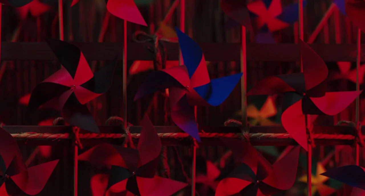 No Longer Human (2019) movie still: closeup — A dark wooden grid fence is covered with numerous plastic colorful pinwheels. Red light casts deep…