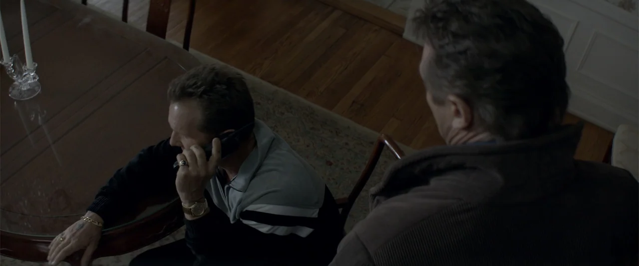 A Walk Among the Tombstones (2014) movie still: medium — A dining room features a round glass-topped table set on a wooden floor. One man sits at…