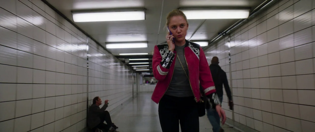 Greta movie still: medium — A long, narrow pedestrian tunnel is lined with white rectangular ceramic tiles. A young woman walks toward the camera…