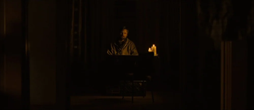 The Road (2009) movie still: wide — A man sits at a table in an almost entirely dark, featureless interior space. The only visible light comes from…