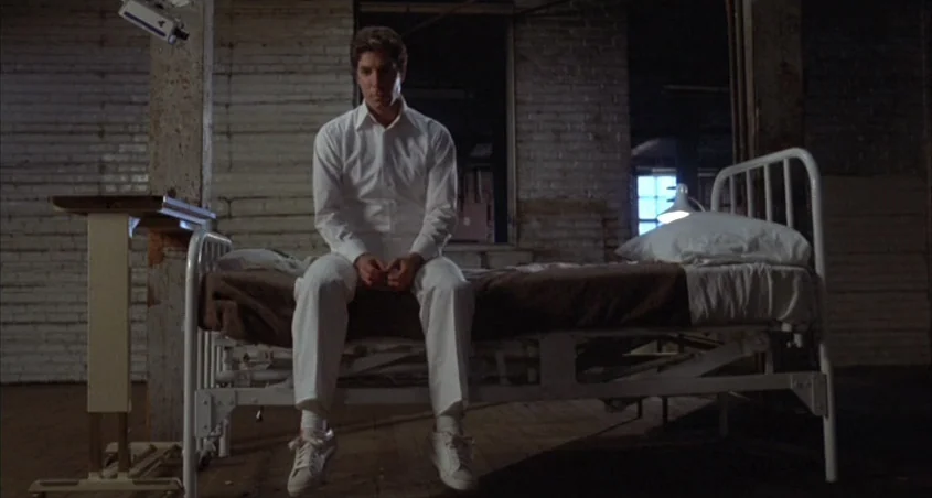 Scanners (1981) movie still: fullbody — A room features white painted brick walls and an old metal frame bed. A man in white clothing sits on the…