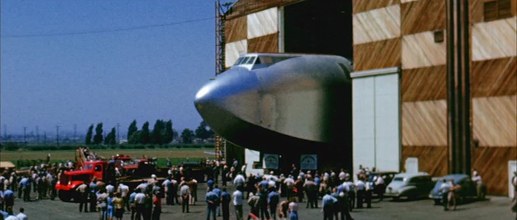 The Aviator (2004) movie still: wide — A crowd of people stands on a tarmac outside a massive wood-sided airplane hangar. The nose of a large silver…
