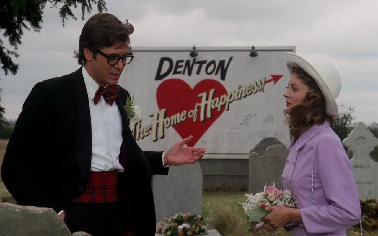The Rocky Horror Picture Show (1975) movie still: medium — A cemetery featuring weathered tombstones sits in front of a large, painted welcome sign.…