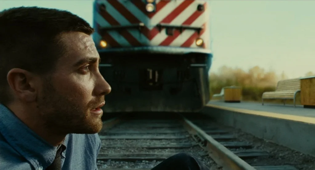 Source Code (2011) movie still: medium — A man sits on railway tracks at a suburban train stop. A commuter train approaches along the tracks in the…