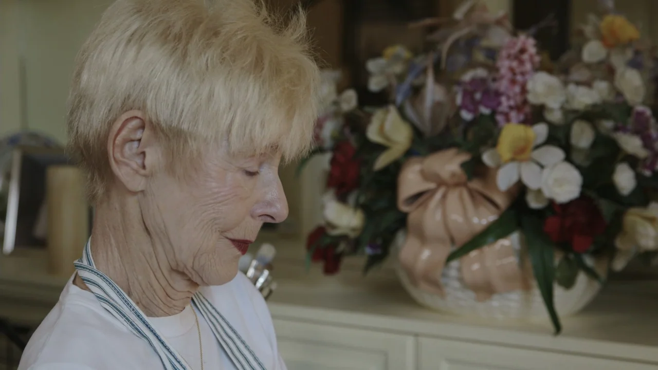 The Last Race (2022) movie still: closeup — An interior space features a floral arrangement in a decorative basket on a shelf. A woman with short…