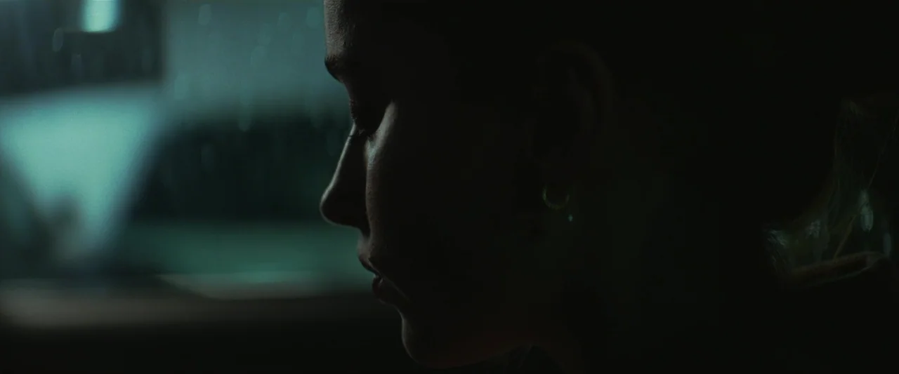 Fair Play (2023) movie still: closeup — A dark, underexposed room holds a person in deep shadow. Only the side profile of a woman's face is…