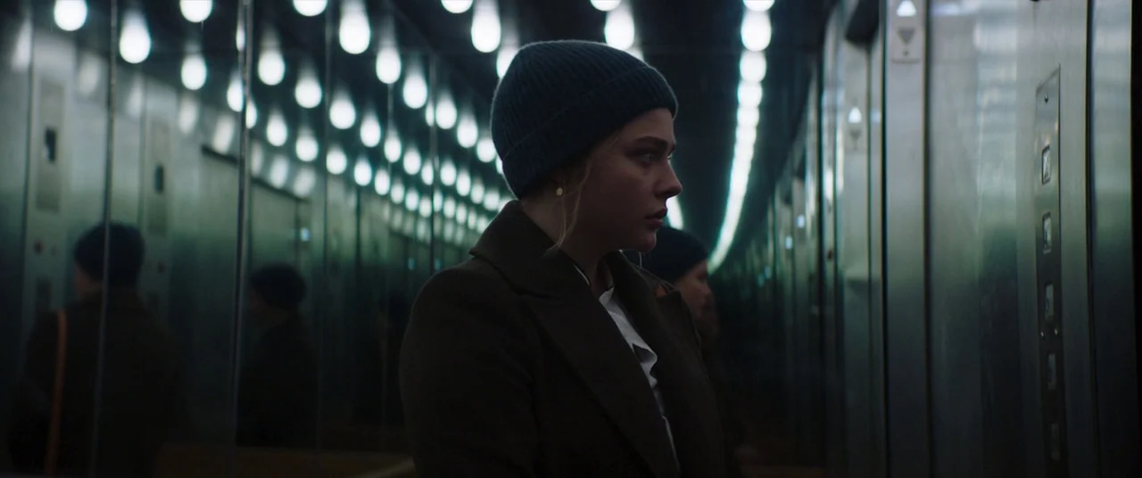 Greta movie still: medium — A polished modern elevator with mirrored panels that create a repeating infinite reflection. A woman in a wool hat and…