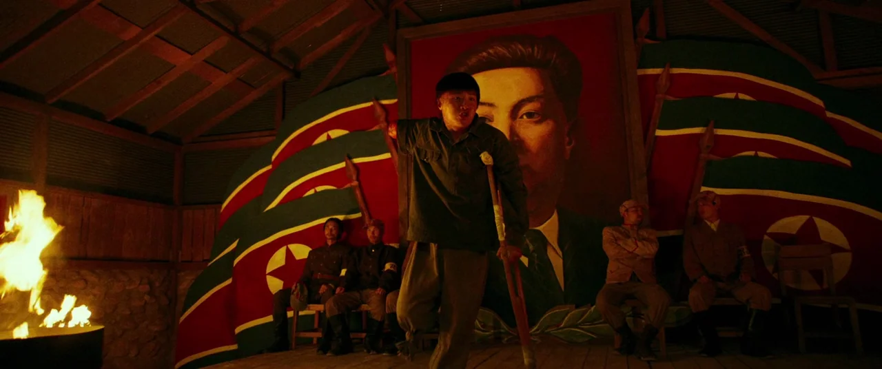 Swing Kids (2018) movie still: fullbody — A wooden shack features a large portrait of a leader against a red wall. Several men sit on benches in the…