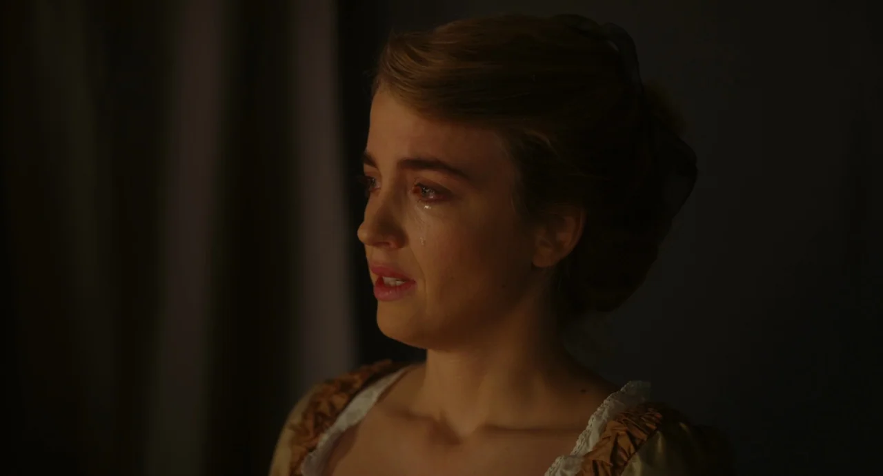 Portrait of a Lady on Fire movie still: closeup — A dimly lit room provides a plain, dark background for a woman in historical clothing. A woman…
