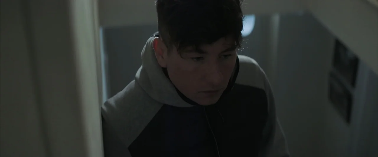 Bring Them Down (2025) movie still: medium — A dimly lit residential stairwell with plain walls and shadow-filled corners. A young man walks down…