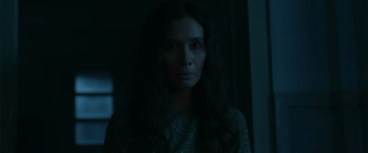 The Unholy (2022) movie still: medium — A dimly lit interior corridor is visible in a cool, desaturated blue color palette. A woman with long dark…