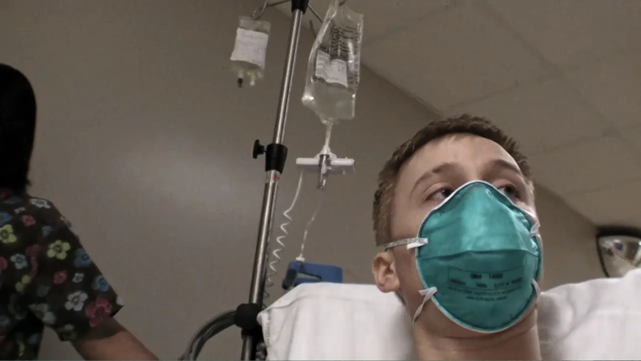 Daniel (1983) movie still: medium — A hospital room contains a patient in a bed and an IV pole with bags. A young man wearing a turquoise mask lies…