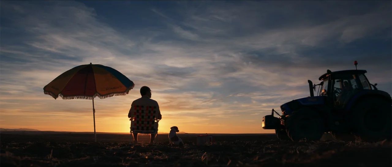 Historias lamentables (2021) movie still: wide — An open plowed field sits under a vast sky at sunset. A man and a small dog sit on folding chairs…