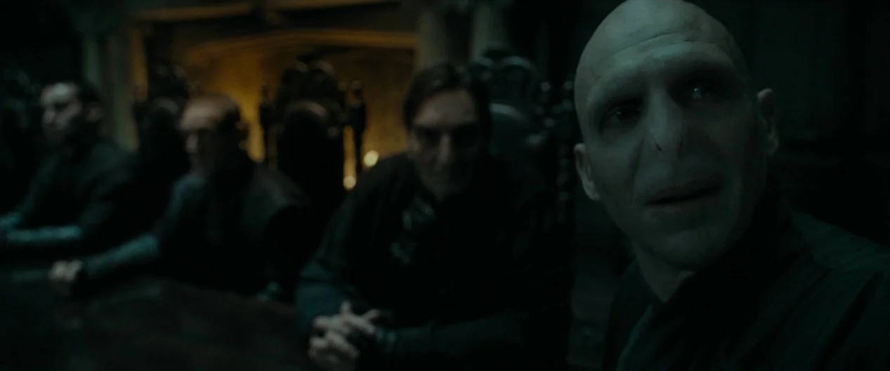Harry Potter & The Deathly Hallows Part 1 (2010) movie still: medium — A dimly lit, stone-walled chamber contains a long table surrounded by seated…