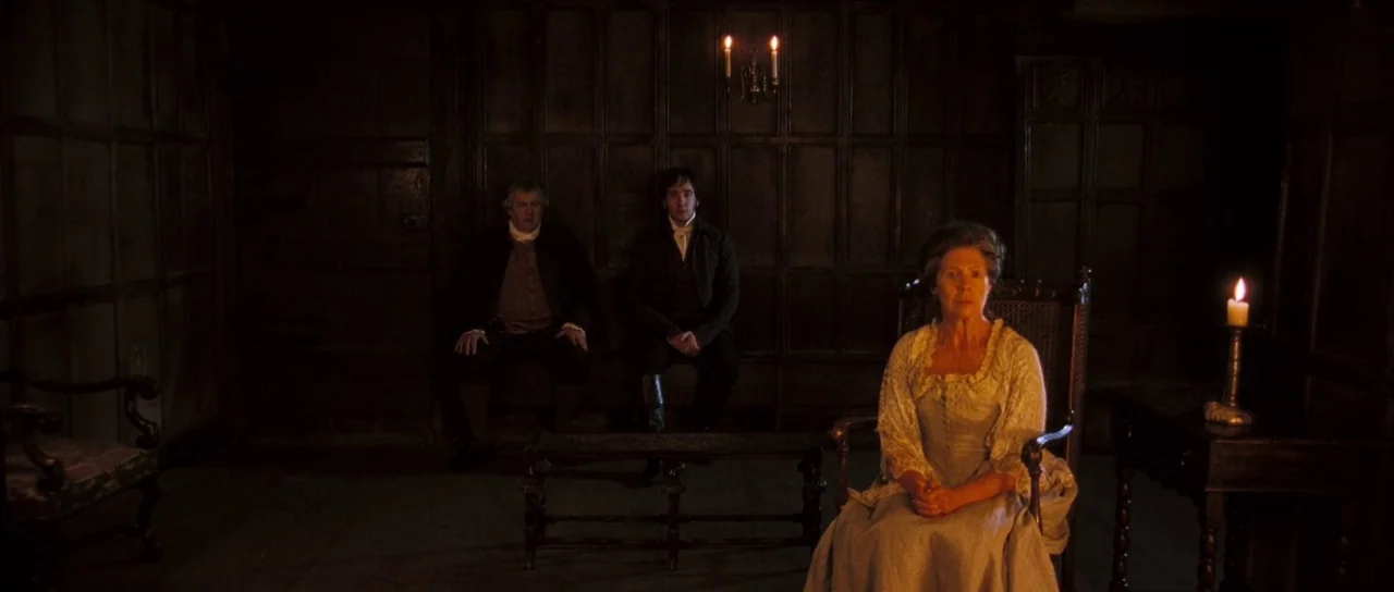 Pride & Prejudice (2005) movie still: wide — A dark, wood-paneled room features dark oak walls and sparse period furniture. Three figures sit in…