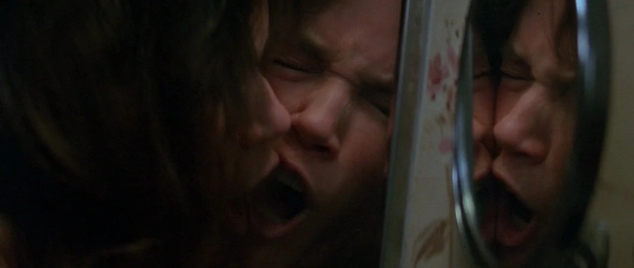 The Entity (1982) movie still: closeup — A dimly lit bathroom features a wall-mounted mirror with a floral patterned frame. A young man yawns with…