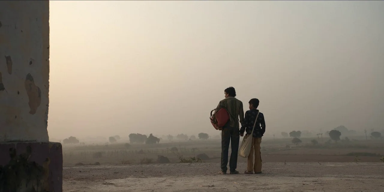 The White Tiger (2021) movie still: fullbody — An open rural road stretches into a hazy, mist-covered agricultural field at sunrise. Two individuals…