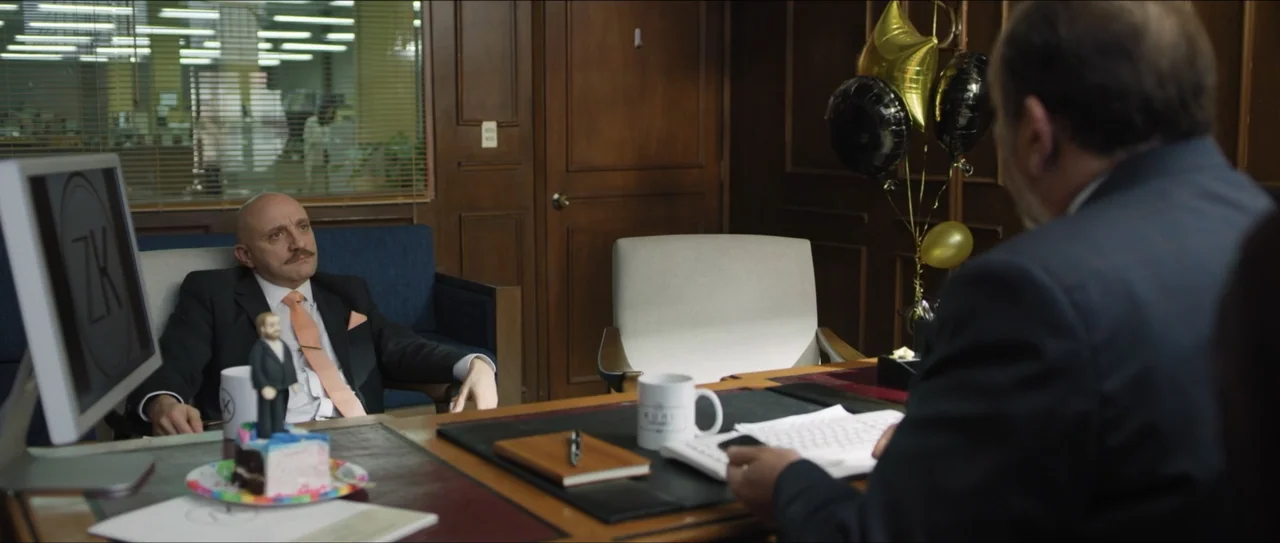 Mirreyes vs Godínez (2019) movie still: medium — A corporate office with wooden paneling, party balloons, and a desk with a monitor. A man in a suit…