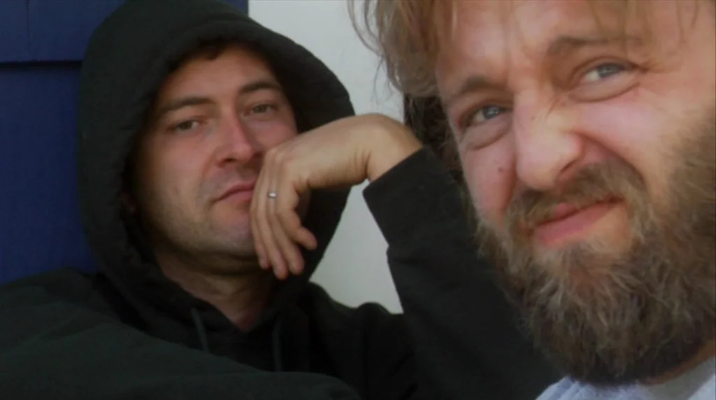 Humpday (2009) movie still: closeup — Two men lean against a blue-painted exterior wall of a house. The men look into the camera while positioned…