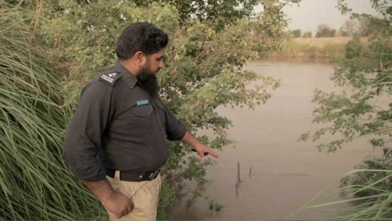 A Girl in the River: The Price of Forgiveness (2015) movie still: medium — A man stands on a muddy bank beside a slow-moving river surrounded by…