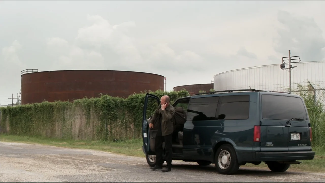 Parker (2013) movie still: wide — An outdoor area features large rusted circular metal tanks behind a vine-covered chain-link fence. A bald man in a…