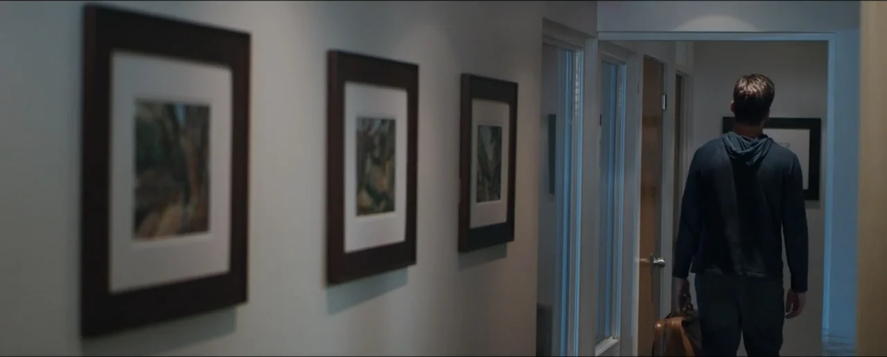 Hostages (2017) movie still: fullbody — A modern home hallway features light-colored walls adorned with three framed landscape paintings. A man…