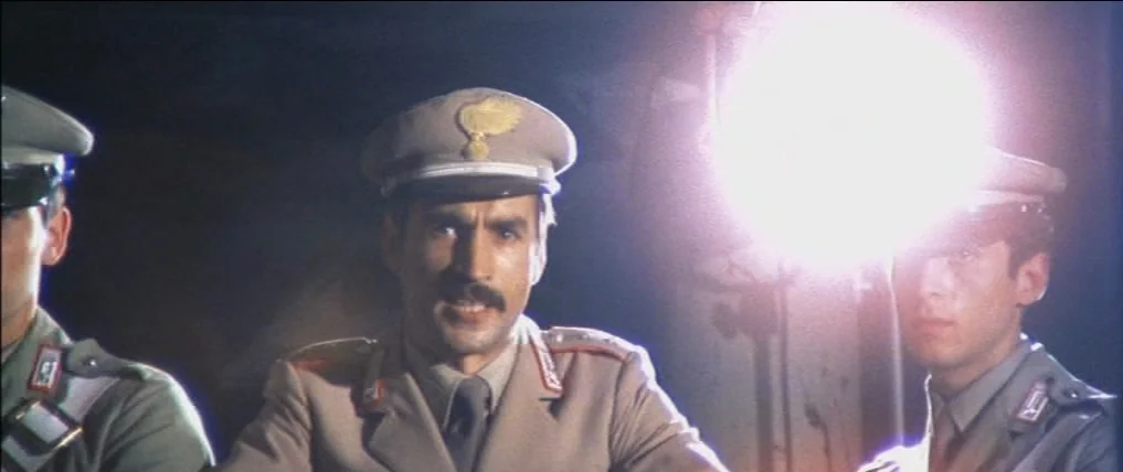 Don’t Torture A Duckling (1972) movie still: medium — Three men in military uniforms stand in the darkness at a nighttime location. A bright light…