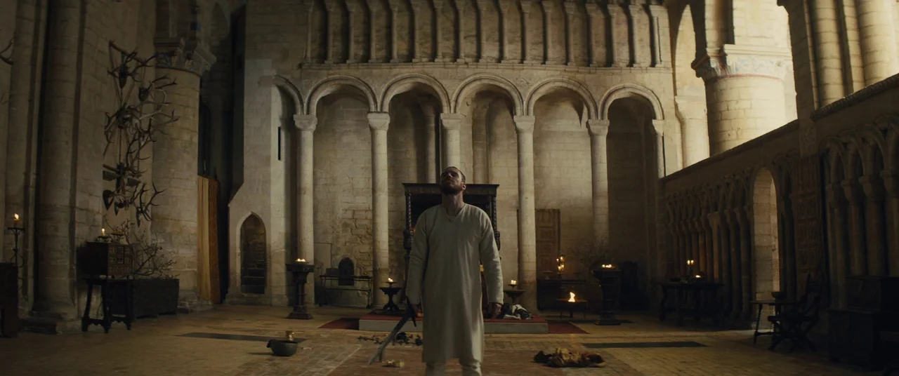 Macbeth (2015) movie still: fullbody — A spacious medieval stone hall features high arched columns and elaborate wall-mounted animal antlers. A lone…