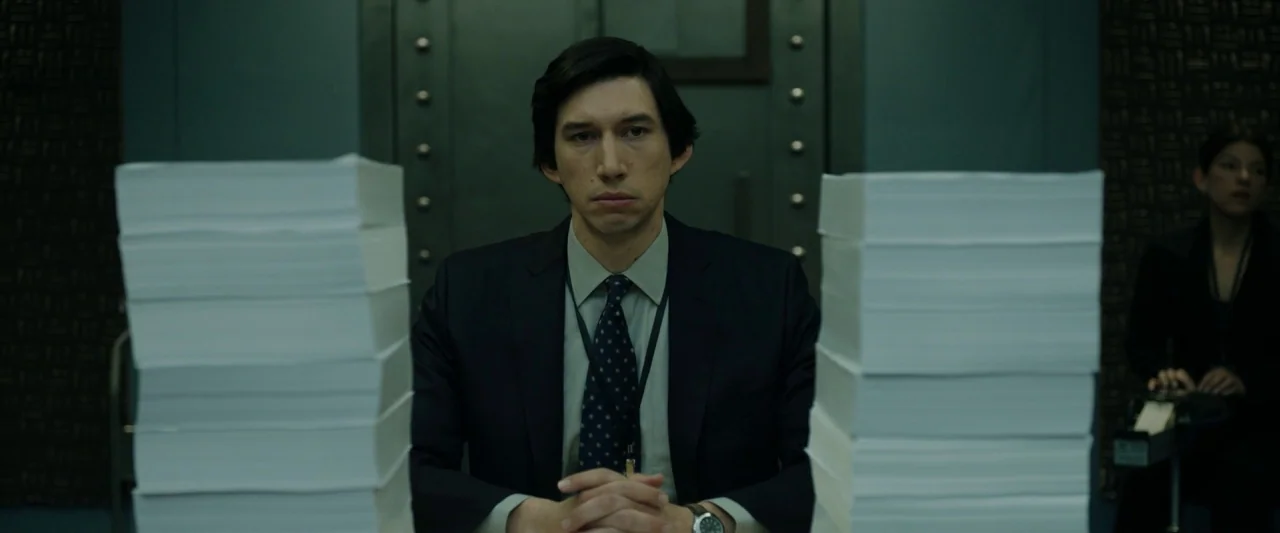 The Report (2019) movie still: medium — A man sits in a windowless room with a heavy reinforced door in the background. Two identical stacks of…