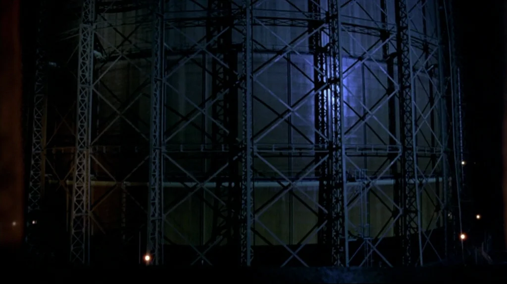 Spider (2002) movie still: establishing — A massive metal gas holder structure stands against a dark night sky. The industrial iron framework…