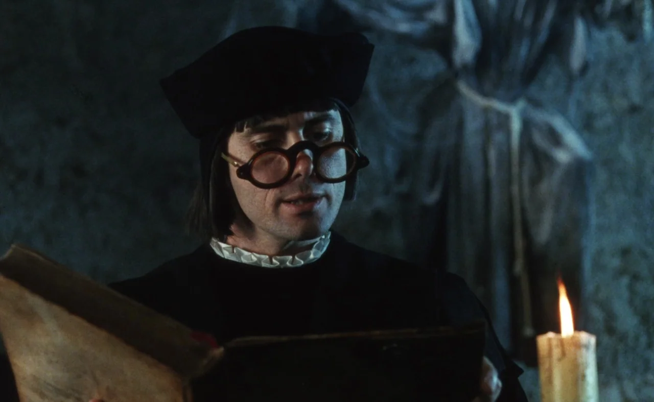 The Pit and The Pendulum (1991) movie still: closeup — A dim, textured stone room serves as the backdrop for a reading setting. A man in period…