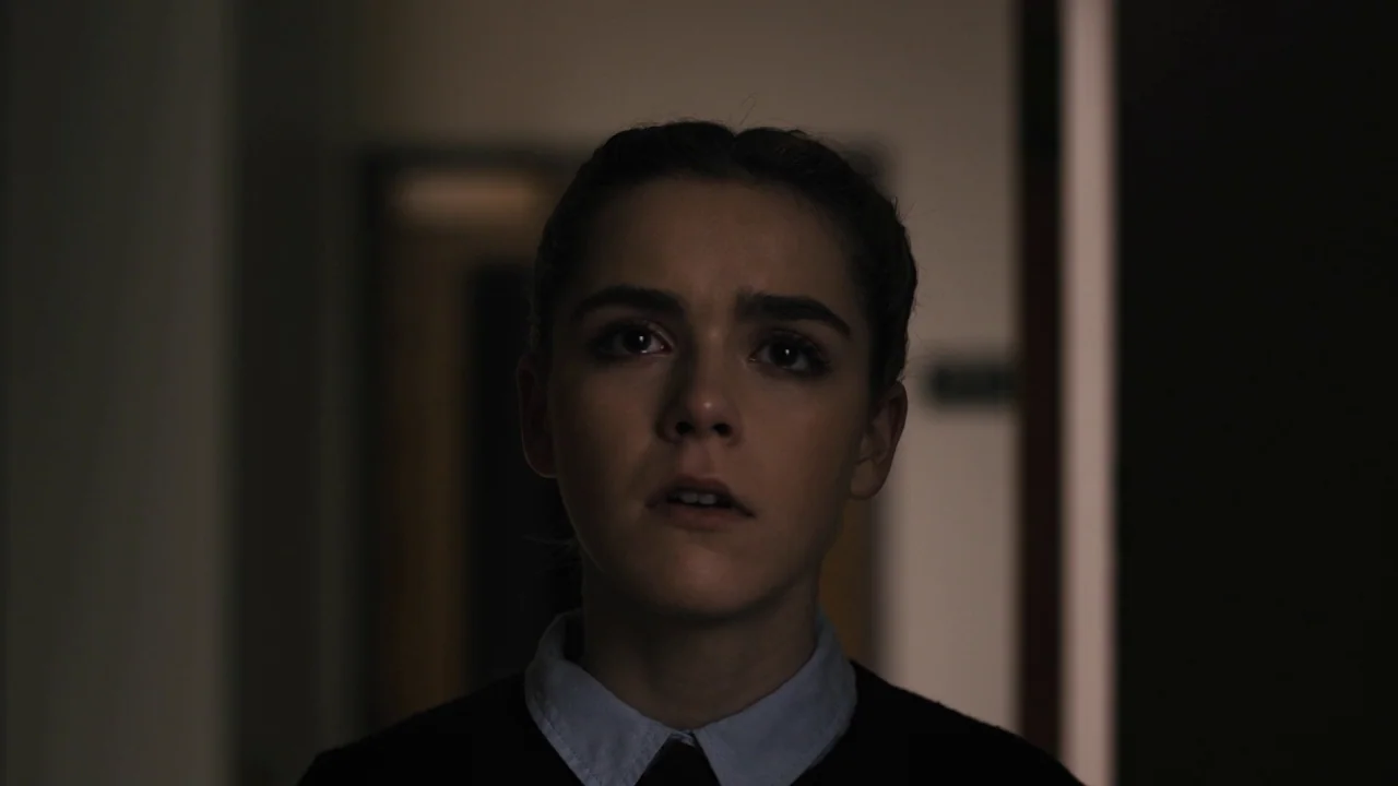 The Blackcoat’s Daughter (2015) movie still: closeup — A dimly lit narrow hallway with blurred architectural features in the background. A young…