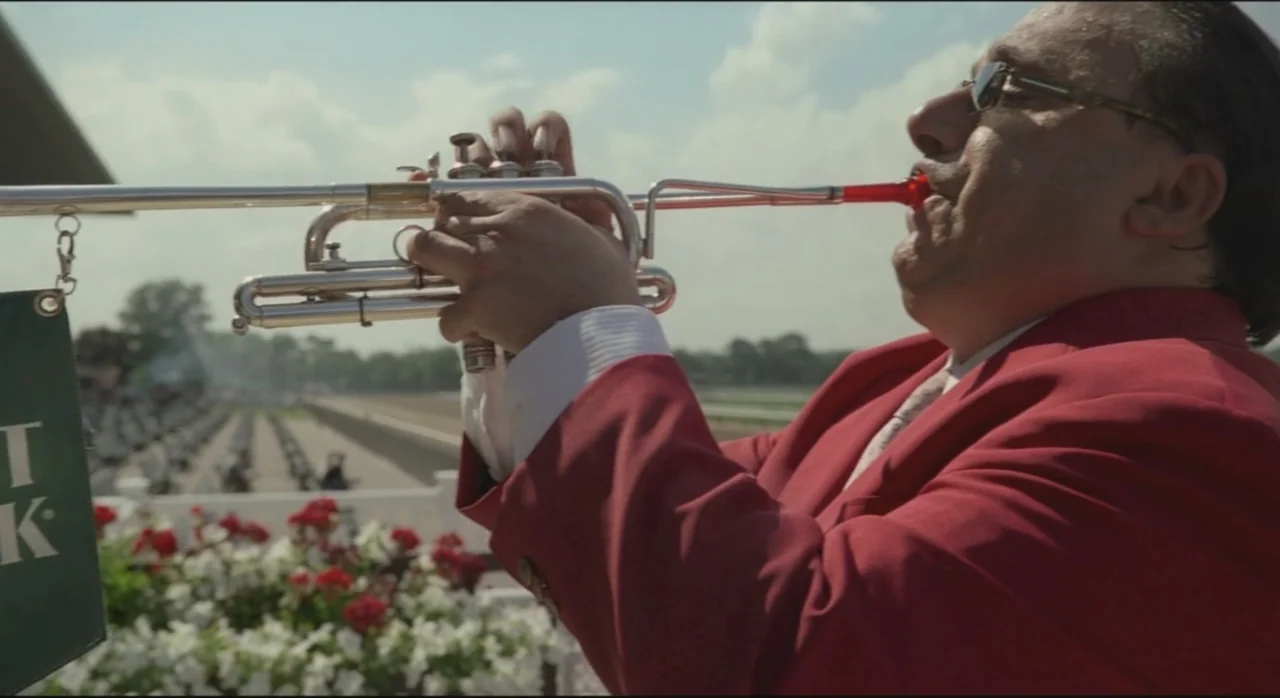 St. Vincent (2014) movie still: medium — A man in a red blazer stands outdoors on a racetrack grounds. He holds a silver trumpet to his lips while…