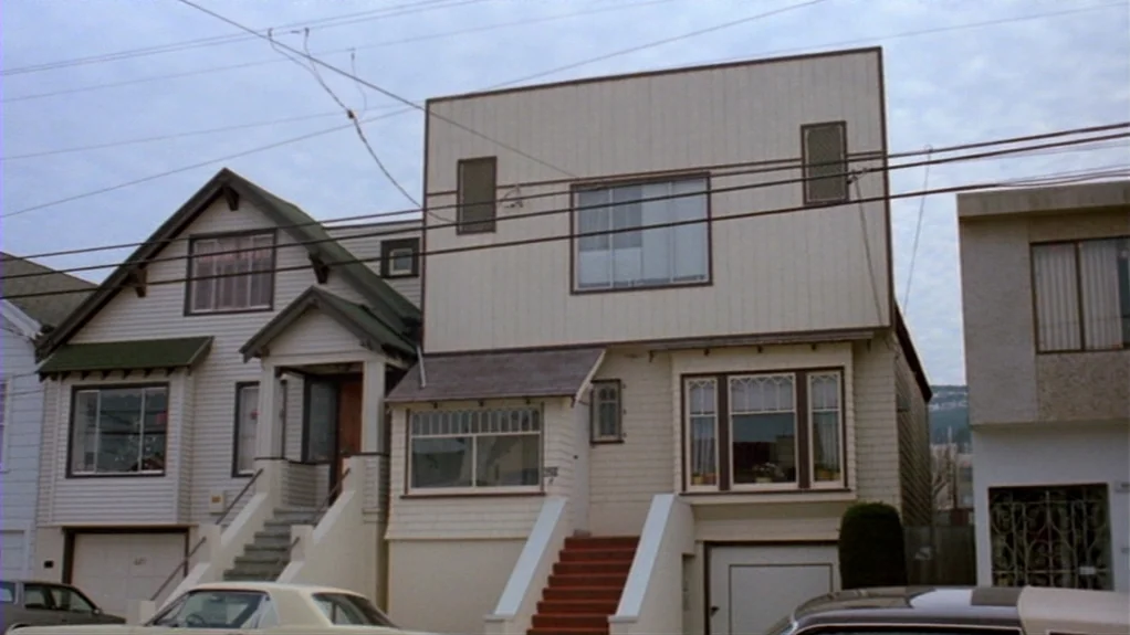 Patty Hearst (1988) movie still: establishing — A residential street in San Francisco features multi-story houses with white siding and porches.…