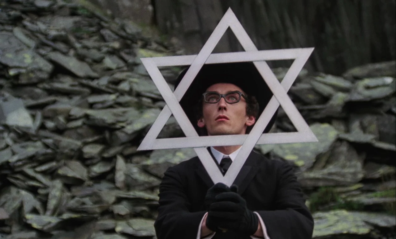 Mahler (1974) movie still: medium — A man stands before a pile of jagged grey stones in an open area. He holds a large silver Star of David directly…