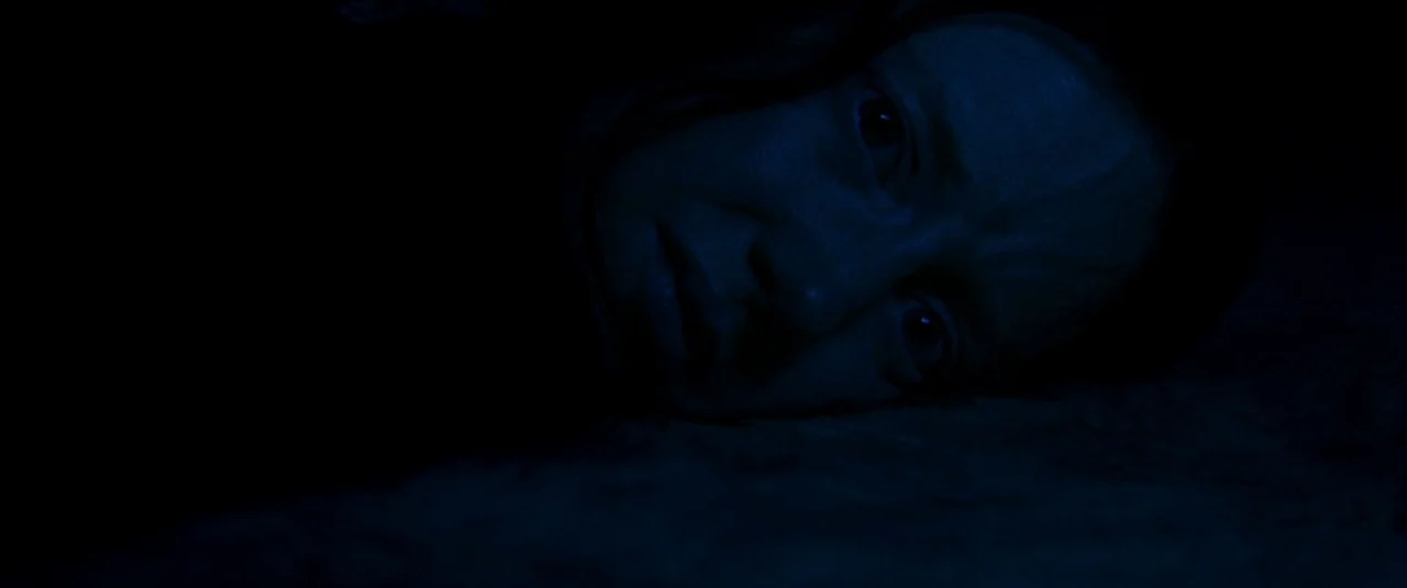 Mandy (2018) movie still: closeup — A dark, indistinct space with a textured, low-lit surface in the foreground. A person lies on the ground with…