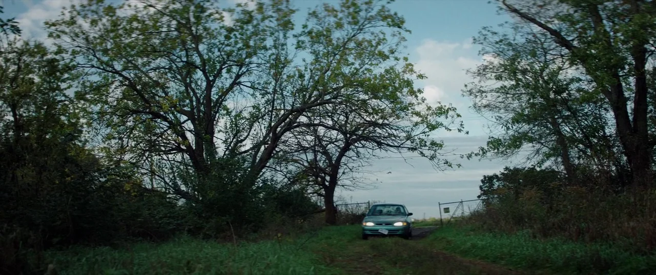 Broadcast Signal Intrusion (2021) movie still: wide — A rural dirt path winds through dense trees and overgrown tall grass. A small passenger car…
