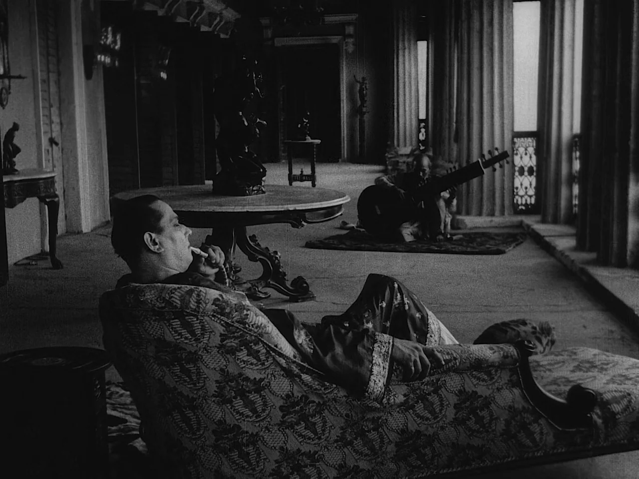 The Music Room (1958) movie still: medium — A spacious, dimly lit room features marble floors and long curtains beside tall windows. A man reclines…