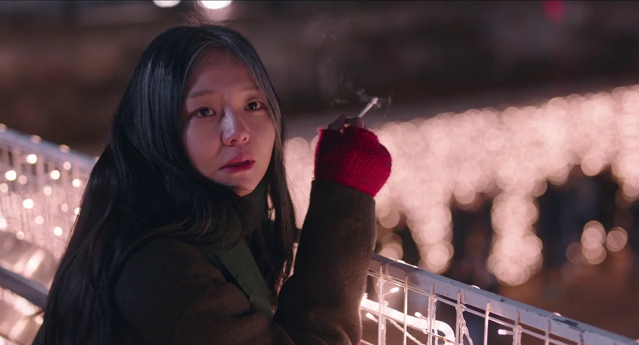 Microhabitat (2017) movie still: closeup — A woman stands on a footbridge decorated with white string lights at night. She holds a lit cigarette…