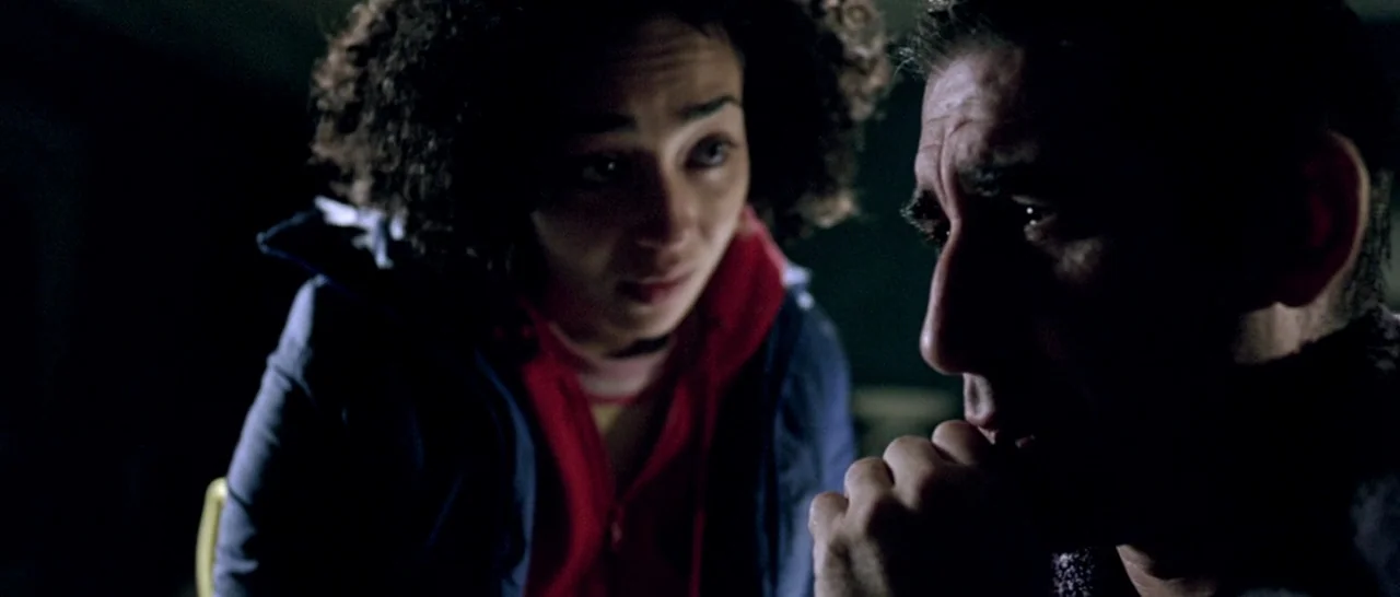 Isolation (2005) movie still: medium — A dimly lit, shadowed space provides the setting for two people sitting close together. A man rests his hand…