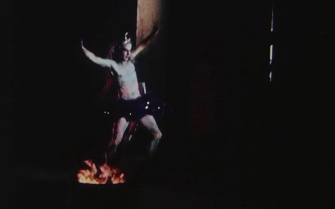 The Last of England (1987) movie still: fullbody — A dark, undefined interior space is dominated by the flickering light of a fire. A person dressed…