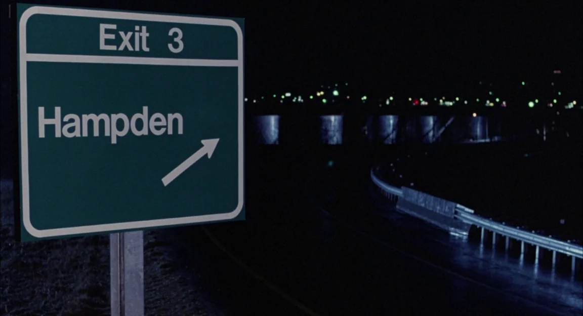Creepshow 2 (1987) movie still: medium — A green highway exit sign stands in the foreground beside a dark asphalt road. Distant city lights twinkle…