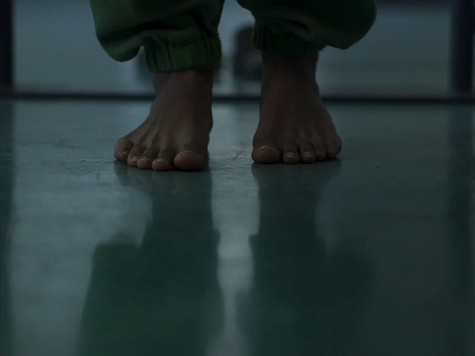 Mogul Mowgli (2020) movie still: closeup — A person stands barefoot on a smooth, slightly reflective floor surface. Their legs are framed from the…