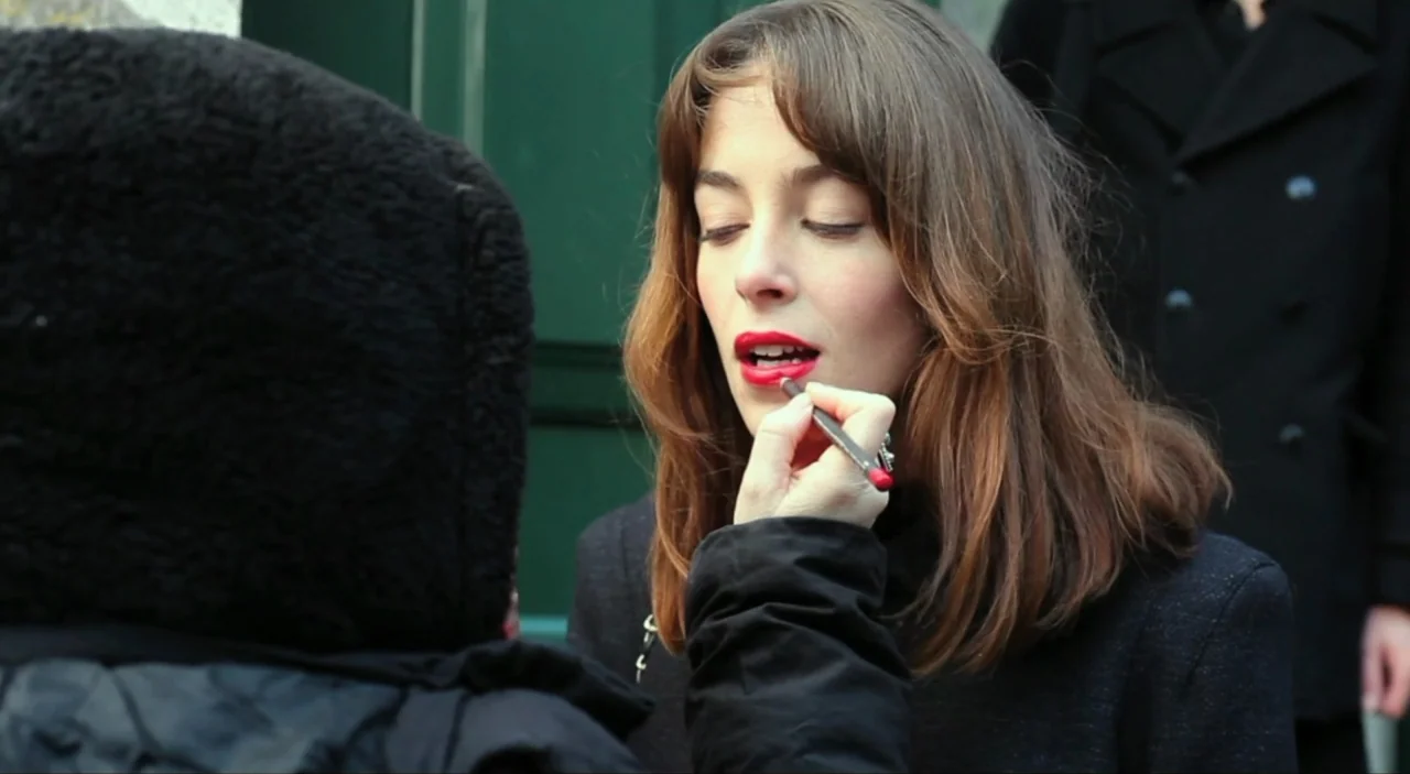 Cosmos (2015) movie still: medium — A woman in a dark coat sits outdoors with a green door behind her. A person in a black hoodie applies bright red…