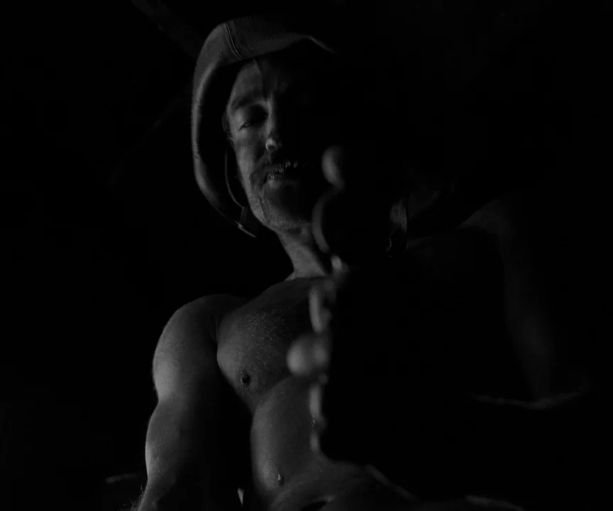 The Lighthouse (2019) movie still: medium — A man stands in darkness wearing a hat while shirtless in a dim space. He reaches his arm out toward the…