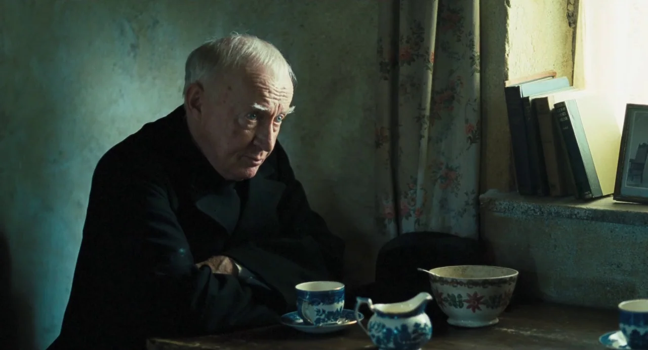 Jimmy’s Hall (2014) movie still: medium — An older man sits at a wooden table in a dimly lit room with plain walls. He rests his arms on the surface…