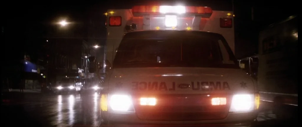 Bringing Out The Dead (1999) movie still: medium — An ambulance drives down a dark urban street at night with headlights illuminating the road. The…