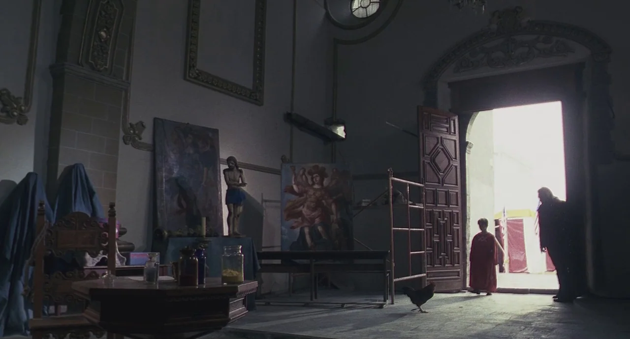 Santa Sangre (1989) movie still: wide — An interior of a church with religious paintings and statues under dim lighting conditions. A child in a red…