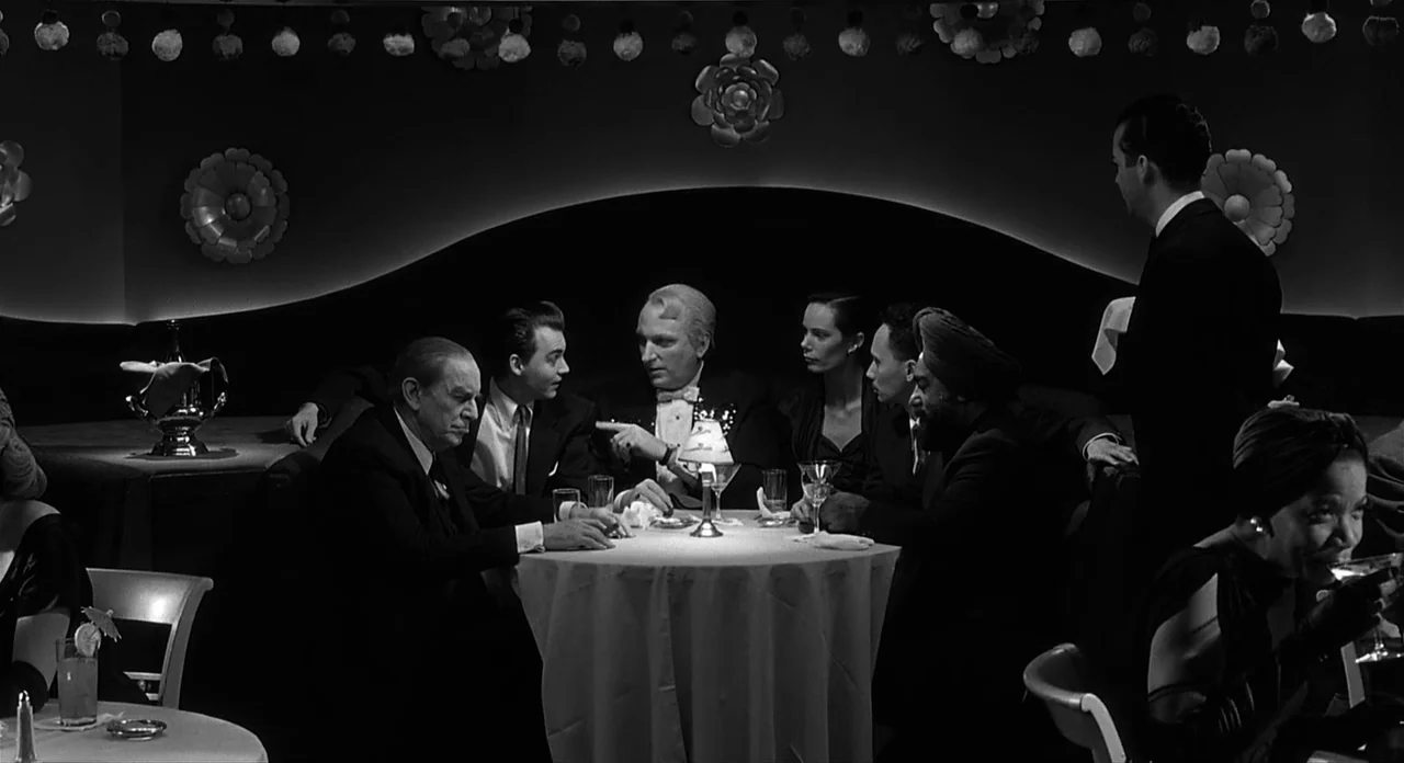 Ed Wood (1994) movie still: wide — A dimly lit club with a circular table where several people in formal wear gather. A man in a tuxedo gestures…